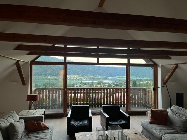 Blick aus dem Wohnraum durch die Panoramaverglasung über den Balkon auf den Ossiacher See und die Berge
